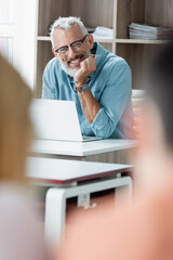 Smiling mature teacher sitting near laptop in school