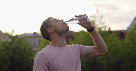 Medium shot of thirsty young man drinking water in plastic bottle outdoors.