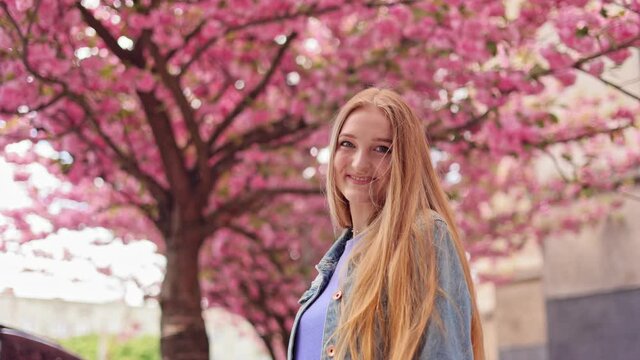 Portrait shot of attractive young woman looking and smiling at the camera on blurred background in the city street. Sacura tree. Close up. Happy Caucasian girl outdoor.