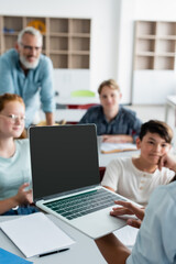 Fototapeta premium Schoolboy holding laptop near blurred multiethnic classmates and teacher