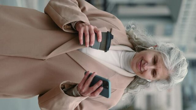 Vertical video. Mature business woman with earphones and mobile on street