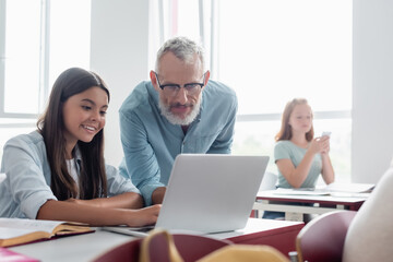 Teacher looking at laptop near smiling schoolgirl in classroom