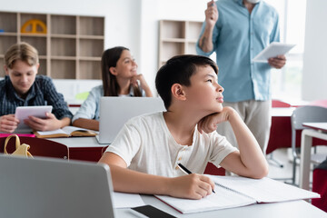 Asian schoolboy looking away while writing on notebook near laptop in classroom