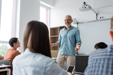 positive teacher gesturing while speaking near teenagers during lesson in school