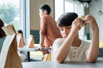 sad asian boy holding sandwich while sitting alone in school canteen near blurred pupils