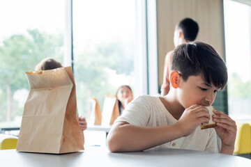 asian schoolboy eating sandwich alone in dining room near blurred teenagers