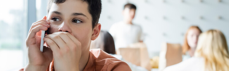 schoolboy covering mouth with hand while talking on mobile phone near blurred classmates, banner