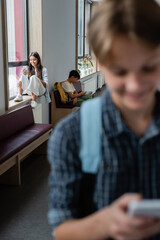 blurred teenager with smartphone near classmates using gadgets on windowsill and bench in corridor