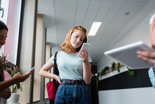 redhead girl looking at mobile phone near blurred teenagers with gadgets
