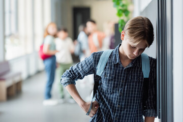 upset schoolboy standing alone with bowed head near blurred pupils in school corridor