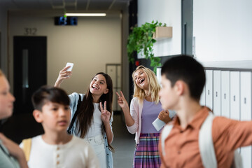 cheerful girls showing victory gesture while taking selfie near blurred classmates