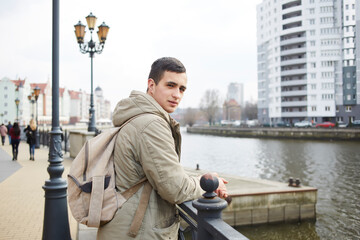 Half length portrait of handsome Caucaisan tourist with casual backpack posing at city embankment, young hipster guy 20s looking at camera during travel vacations for visiting town with quay