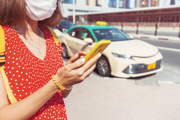 Female tourist in a medical protective mask during the Covid pandemic calls a taxi using an application in a smartphone for a trip in the city