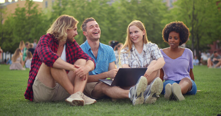 Happy young diverse friends smiling and browsing laptop while sitting on grass in park together