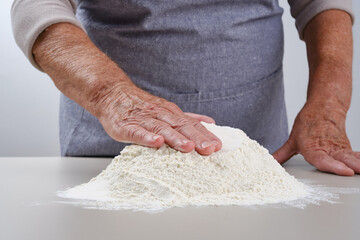 Female hands of an elderly woman with a heap of flour on a gray table. Older woman in a gray apron preparation of dough for a pizza, bread, pasta, festive cake. Grandmother making pasta at home