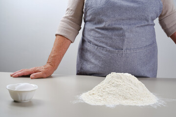 Elderly woman with a heap of flour and egg on a gray table. Older woman in a gray apron preparation of dough for a pizza, bread, pasta, cake. Grandmother making pie at home. Focus on a heap of flour