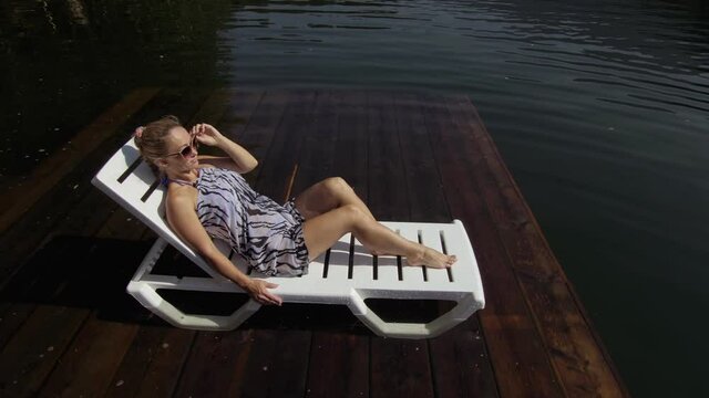 Woman lie on a sunbed in sunglasses and a boho silk shawl. Girl rest on a flood wood underwater pier. The pavement is covered with water in the lake. In the background are mountain and a forest.