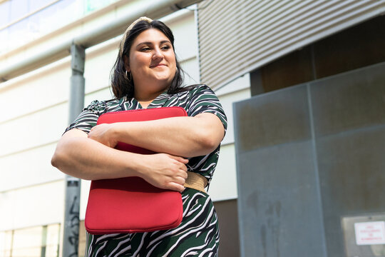 Happy woman with laptop case standing near urban building