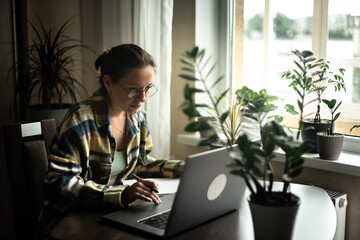 Focused woman in domestic wear and eyeglasses sitting near the window at cozy workplace and...