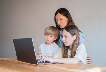 Fototapeta na wymiar Mom with two children are sitting at home at the table with a laptop. Distance learning of children. Mom helps children learn remotely