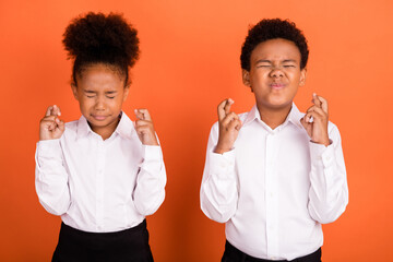 Photo of two young afro school kids crossed fingers worried expect exam wish luck isolated over orange color background