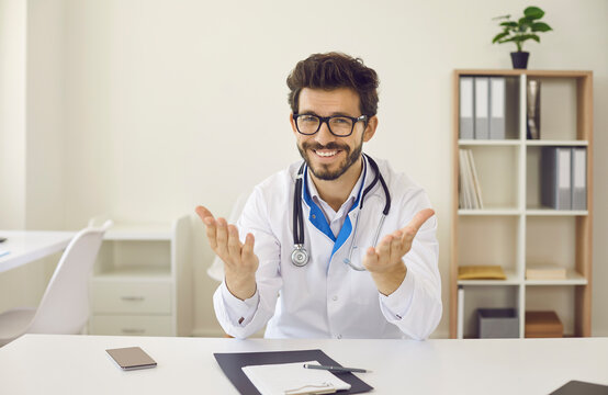 Happy handsome male doctor in white coat and glasses sitting at table in medical office, looking at you and talking. Computer device video call screen headshot for online discussion with patient