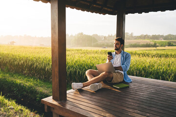 Young bearded man with smartphone and netbook working in gazebo on meadow