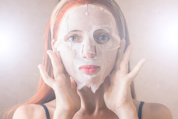 A redhead girl with a bandage on her head applies a fabric white nourishing mask to her face and looks at the camera.