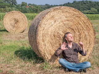 Naklejka na ściany i meble A middle-aged white man listens to music on headphones sitting in a field, with his back to a bale of hay