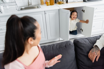 cheerful asian kid hiding in kitchen cabinet near blurred parents on sofa