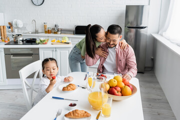 asian wife kissing husband near daughter having breakfast in kitchen