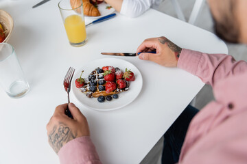 cropped view of tattooed man holding cutlery near waffles with berries on plate