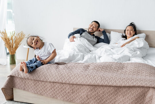 Cheerful Asian Kid Sitting On Bed Near Blurred Parents