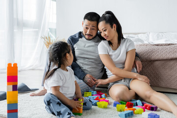 Obraz premium cheerful asian husband and wife holding hands and looking at toddler daughter playing building blocks
