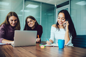 Cheerful Asian woman with education textbook for planning organization calling via cellphone technology while successful colleagues using laptop for informative research and online banking