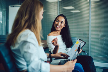 Funny Asian and Caucasian colleagues discussing organization plan during brainstorming in office interior, joyful employees with netbook technology and paperwork report analyzing business strategy