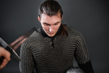 A medieval warrior in chain mail sits on his knee with a helmet in his hands. Knighting.