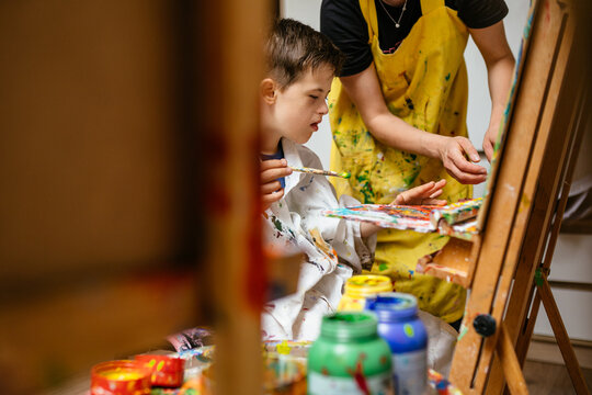 Young Caucasian down syndrome boy sitting at chair with colors and painting on easel with concentrated and calm painting. Concept for education for disabled kid.