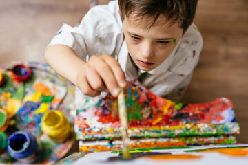 Young Caucasian down syndrome boy sitting at chair with colors and painting on easel with...