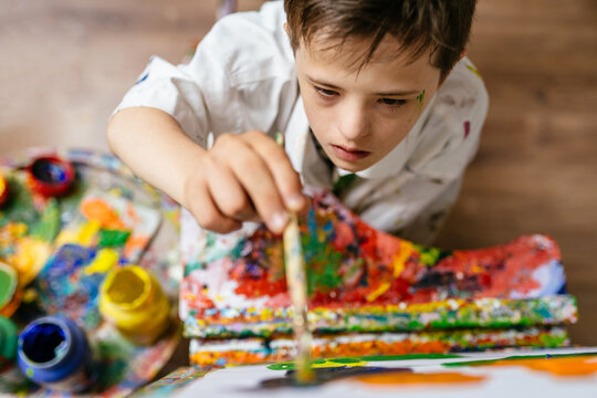 Young Caucasian down syndrome boy sitting at chair with colors and painting on easel with concentrated and calm painting. Concept for education for disabled kid.