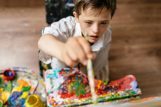 Young Caucasian down syndrome boy sitting at chair with colors and painting on easel with concentrated and calm painting. Concept for education for disabled kid.