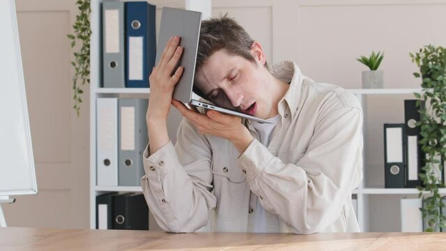 Tired funny sleepy overworked dozing caucasian man millennial businessman male manager user sitting at table in office holding laptop as pillow near ear sleeping dreaming yawning resting at workplace