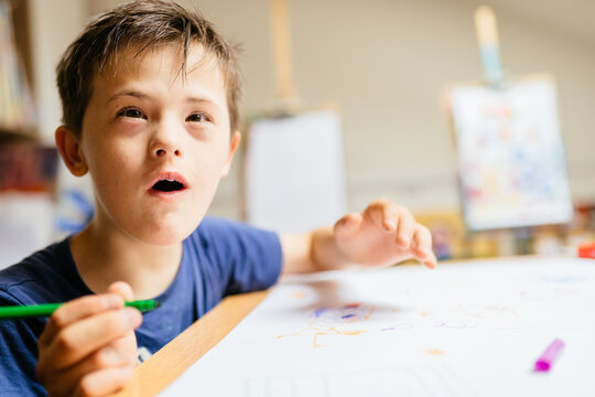 Positive emotions brown hared boy with Down syndrome draw at a table on a white paper at painting studio for special need children