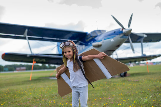 A little girl plays a pilot on the background of a small plane with a propeller. A child in a suit with cardboard wings dreams of flying
