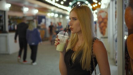 A young girl drinks a refreshing cocktail with a straw, in the evening, on a street lit by lights. Rest in a street cafe.