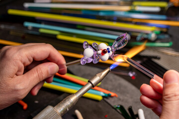 Hands of the glassmaker, making figurines from glass. Shaping hot murano glass into a bunny under a jet of fire from a gas burner.  Handmade fine art. Selective focus. Close up. - obrazy, fototapety, plakaty