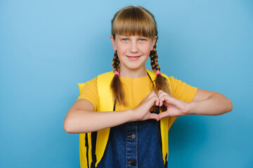 Adorable cute little schoolgirl showing heart gesture with hands, smiling looking at camera, wears yellow backpack, posing isolated over blue color background in studio. Back to school concept