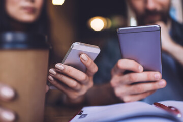 Cropped image of male and female hands using modern cellphone devices for online communication during organisation planning in coffee shop, selective focus on digital smartphones and takeaway cup