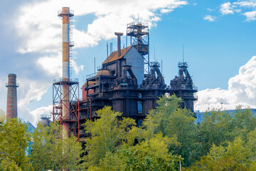 Fototapeta na wymiar The building of the old factory on a background of blue sky with clouds. Metallurgical plant pipes. Iron smelter behind the tree line. Black storage containers.