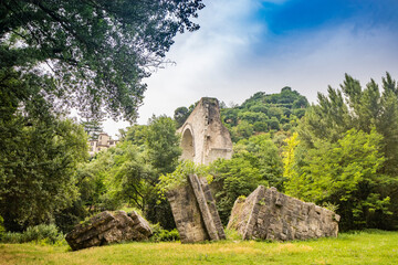 The ruins of the Roman arch bridge of Augustus, in Narni, Terni, Umbria. The remains of the bridge over the Nera river. The big and ancient stone arch, against the blue sky. Trees and dense vegetation - obrazy, fototapety, plakaty